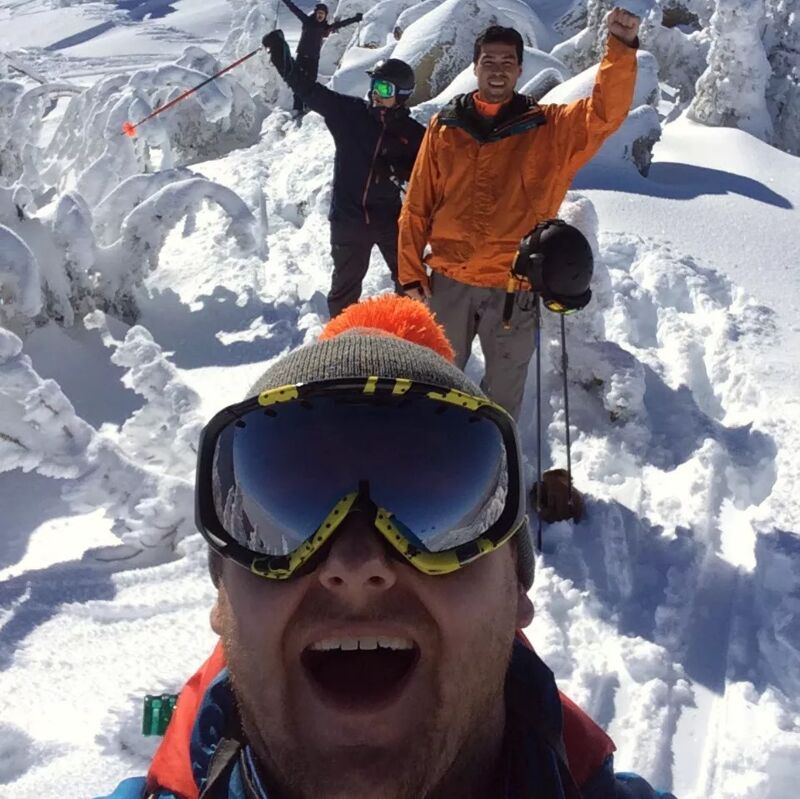 The image shows a group of people in a snowy mountain environment. One person is taking a selfie, while others are in the background, possibly skiing or snowboarding. They appear to be enjoying a winter activity. The overall mood is joyful and adventurous.